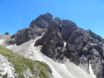 Ritornati al Gera si scende, rasentando a sinistra le pareti della Croda di Tacco. Si nota una traccia tra le ghiaie che pare scomparire per poi risalire su di un costone erboso tramite un’esile cengetta, ove ha inizio la ferrata Mazzetta. Per la precisio